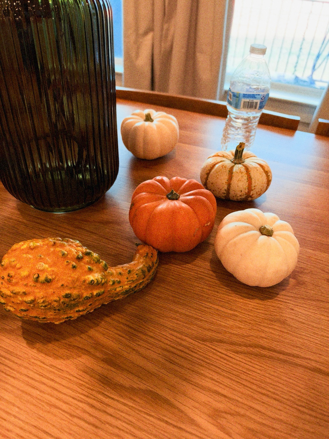 Orange and cream gourds on a wooden table beside a dark green vase, with the mini gourd at the edge.